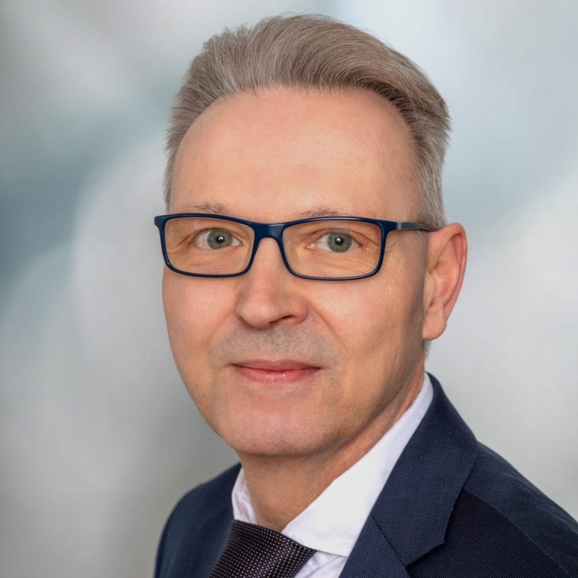 Ein Mann mit Brille, grauen Haaren, in einem Anzug mit Krawatte vor unscharfem Hintergrund.