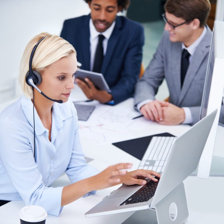 Frau mit Headset arbeitet am Laptop, umgeben von zwei Männern im Office.